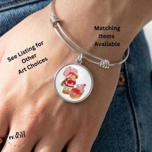 May include: Silver bangle bracelet with a round charm depicting a cartoon character in a pink dress and hat. The charm has a white background. Text on the image says "Matching Items Available" and "See Listing for Other Art Choices".