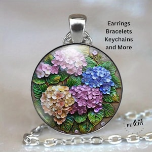 Puede incluir: Un collar con colgante plateado con un diseño floral redondo. El colgante muestra una imagen detallada en relieve de hortensias coloridas en tonos rosa, morado, azul y amarillo, con hojas verdes. El texto "Earrings Bracelets Keychains and More" es visible.