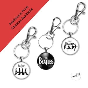 Könnte beinhalten: Schlüsselanhänger mit dem Logo und den Bildern der Beatles. Jeder Schlüsselanhänger hat einen silberfarbenen Verschluss und einen runden Anhänger. Die Anhänger zeigen das Logo der Band, das Albumcover von Abbey Road und eine Silhouette der Bandmitglieder.