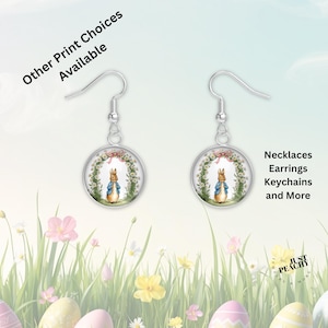 May include: Silver-tone earrings with a circular pendant featuring a Peter Rabbit illustration within a floral arch. The image includes text that reads "Other Print Choices Available" and "Necklaces Earrings Keychains and More". The background has Easter eggs and flowers.