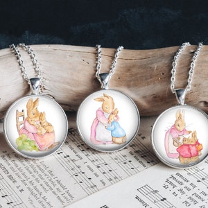 May include: Three silver necklaces with round glass pendants featuring watercolor illustrations of a mother rabbit and her baby bunnies. The pendants are set against a white background.