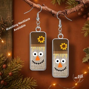 May include: A pair of rectangular dangle earrings featuring a scarecrow face design with a brown hat, a yellow sunflower, a burlap-textured face, white eyes, an orange triangular nose, and a black stitched mouth, hanging from silver-toned hooks.