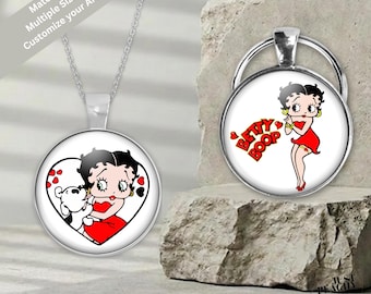 Puede incluir: Un collar con colgante plateado y un llavero circular a juego, ambos con un cabujón de cristal con una ilustración de Betty Boop. El colgante muestra a Betty Boop con un perro dentro de un corazón, mientras que el llavero muestra a Betty Boop con el texto Betty Boop en letras rojas.