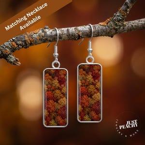 May include: A pair of rectangular drop earrings featuring a vibrant autumn forest canopy print in shades of red, orange, and yellow. The earrings have silver-toned metal frames and hook-style ear wires, displaying a detailed aerial view of trees.
