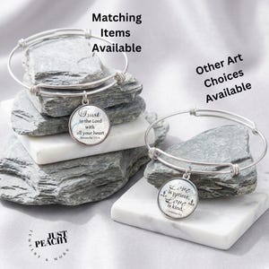 May include: Two silver-tone expandable wire bangles featuring round charms. One charm displays the text Trust in the Lord with all your heart Proverbs 3:5-6. The other charm displays the text Love is patient, Love is kind. 1 Corinthians 13:4. The text Just Peachy Jewelry & More is visible.
