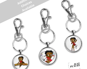 Puede incluir: Tres llaveros redondos con diseños de Betty Boop. Cada llavero tiene un cierre y un anillo de color plateado. Las imágenes de Betty Boop la muestran con un vestido rojo, con un corazón y con las palabras "Betty Boop". El texto "Additional Choices Available" está en el fondo.