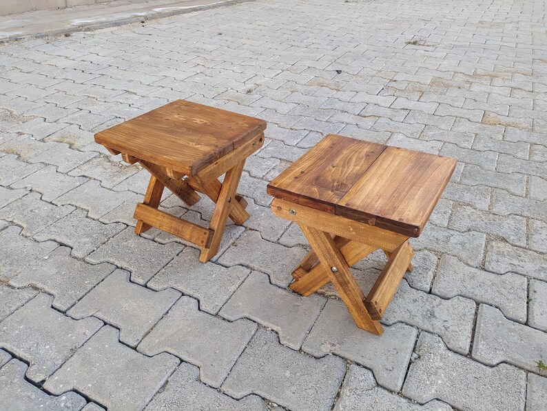 May include: Two wooden folding stools with a natural brown stain finish. The stools are made of solid wood and have a simple, rustic design.