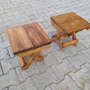 May include: Two wooden folding stools with a natural brown stain finish. The stools are made of solid wood and have a simple, rustic design.