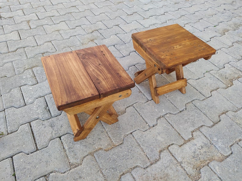 May include: Two wooden folding stools with a natural brown stain finish. The stools are made of solid wood and have a simple, rustic design.