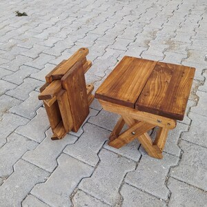 May include: Two wooden folding stools, one is folded and the other is unfolded. The stools are made of natural wood and have a dark brown stain. The stools are sitting on a gray concrete patio.