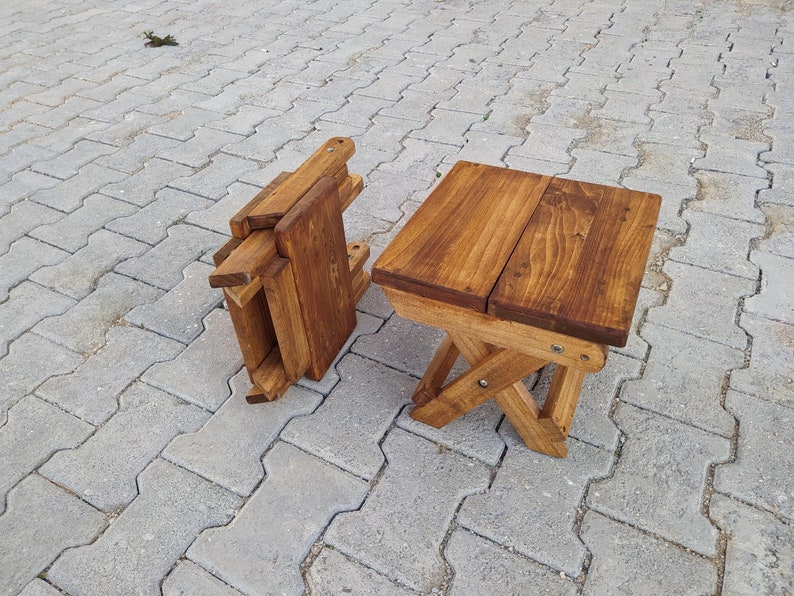 May include: Two wooden folding stools, one is folded and the other is unfolded. The stools are made of natural wood and have a dark brown stain. The stools are sitting on a gray concrete patio.