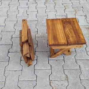 May include: A wooden folding stool with a natural brown finish. The stool is folded up and sitting on a gray concrete surface.