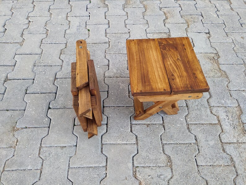 May include: A wooden folding stool with a natural brown finish. The stool is folded up and sitting on a gray concrete surface.
