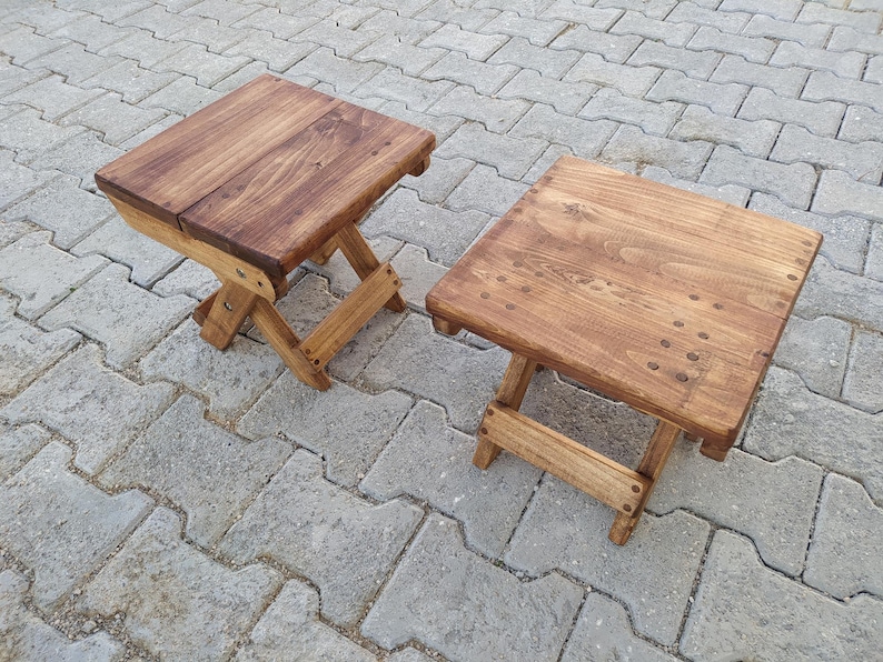 May include: Two wooden folding stools with a natural brown stain. The stools are made of solid wood and have a simple, rustic design.