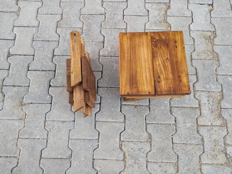May include: A set of two wooden folding stools. The stools are made of natural wood and have a rustic finish. The stools are folded and stacked on top of each other.
