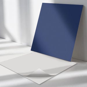 May include: Two square pieces of paper, one white and one blue, are lying on a white surface. The white paper has a corner folded over, revealing a white adhesive backing.