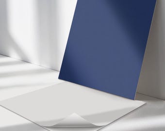 May include: Two square pieces of paper, one white and one blue, are lying on a white surface. The white paper has a corner folded over, revealing a white adhesive backing.