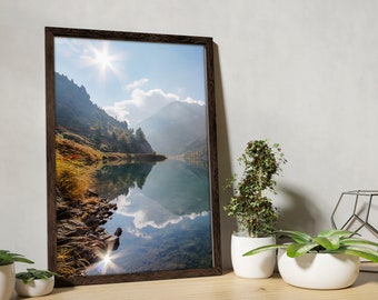May include: A framed landscape photograph of a mountain lake scene. The image features a bright sun, blue sky, and the reflection of the mountains in the water. The frame is dark brown.