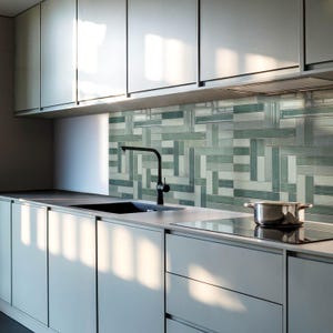 Puede incluir: Diseño de cocina moderno con gabinetes gris claro, un fregadero y grifo negros, y un salpicadero de azulejos de vidrio en tonos verdes y grises. Una olla de acero inoxidable está sobre la estufa. La cocina está bien iluminada.