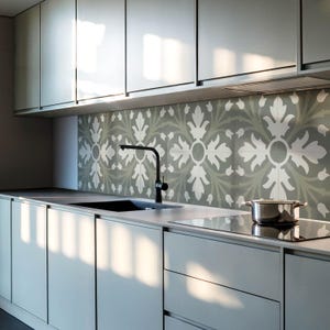 May include: A modern kitchen with white cabinets and a gray tiled backsplash featuring a floral pattern. A black faucet is mounted above a stainless steel sink. A black stovetop is visible in the foreground.