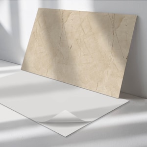 Puede incluir: Una gran losa rectangular de piedra beige con vetas marrón oscuro se apoya contra una pared blanca. Una hoja de papel blanca con una esquina doblada descansa frente a la piedra, creando una composición minimalista.