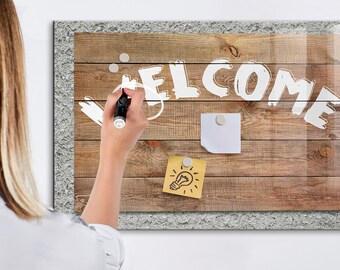 May include: A wooden framed glass dry erase board with a white inscription that says "WELCOME". The board has a light brown wood grain pattern and is decorated with two white push pins and a yellow sticky note with a light bulb drawing. A person is writing on the board with a black marker.