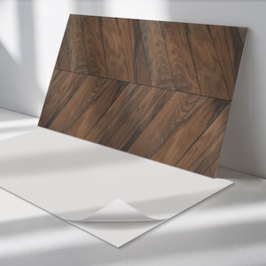 Puede incluir: Una hoja de papel tapiz autoadhesivo con un patrón de veta de madera. El papel tapiz es marrón con un diseño de chevron. La hoja está sobre una superficie blanca con una esquina despegada para mostrar el adhesivo.
