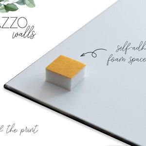May include: The back of a white framed print with a white foam spacer pad attached. The pad has a yellow adhesive backing and the text "self-adhesive foam spacer pads" is written on the white frame. The text "PAZZO walls" is written at the top of the frame.