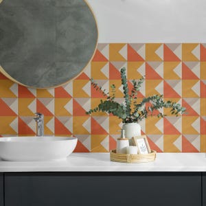 Puede incluir: Un tocador de baño con un lavabo blanco y un espejo redondo. El salpicadero es un patrón geométrico de triángulos naranja, amarillo y beige. Una pequeña planta en un jarrón blanco se encuentra en el mostrador, junto con una bandeja de artículos de tocador.