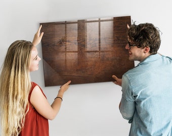 May include: A couple is hanging a brown wood-grain framed picture on a white wall. The picture is glossy and reflects the room.