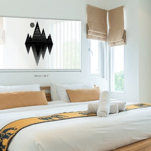 May include: A black and white geometric mountain design print on a white canvas. The print is 100 cm wide and 50 cm tall. The print is hanging on a white wall above a bed with white bedding and a yellow throw blanket.