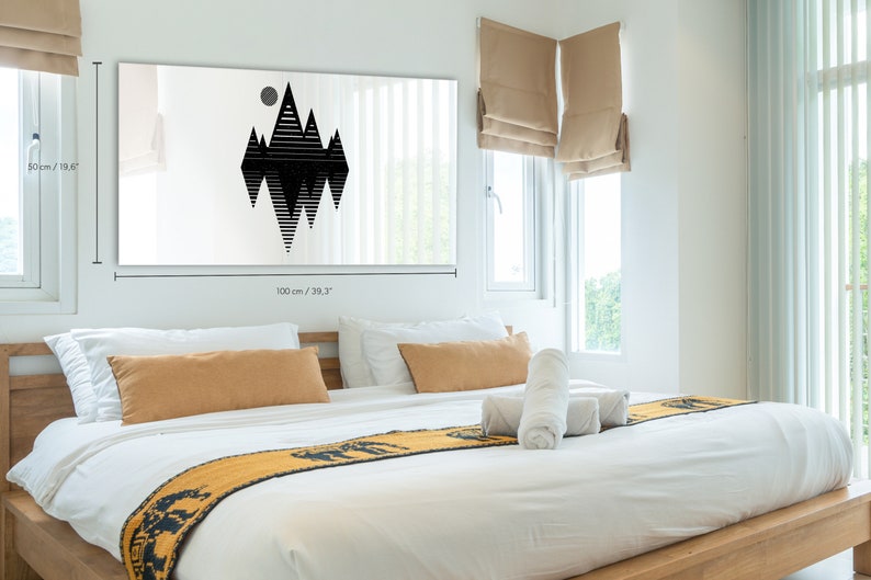 May include: A black and white geometric mountain design print on a white canvas. The print is 100 cm wide and 50 cm tall. The print is hanging on a white wall above a bed with white bedding and a yellow throw blanket.