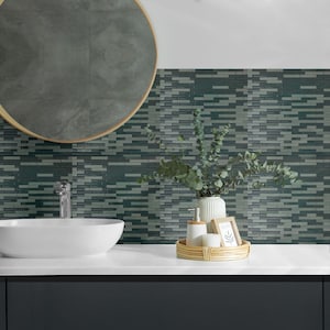 May include: A modern bathroom scene featuring a white oval sink, a round mirror with a gold frame, and a mosaic tile backsplash in shades of teal and gray. A woven tray holds a soap dispenser, soap, and a small framed picture.