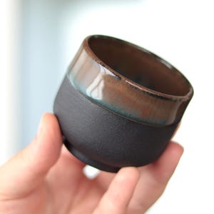 May include: A handmade ceramic cup with a matte black base and a glossy, brown-glazed rim. The rim has a subtle teal accent. The cup is held in a hand, showcasing its unique shape and texture. This piece is ideal for enjoying coffee or tea.