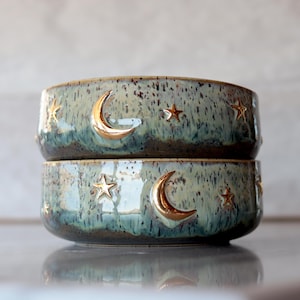 May include: Two ceramic bowls stacked on top of each other. The bowls have a speckled, teal glaze with gold-colored crescent moons and stars. The bowls are round and have a glossy finish.