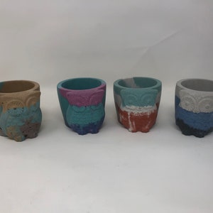 May include: Four small owl-shaped concrete planters in different color combinations. The planters are painted in shades of blue, brown, pink, and gray.