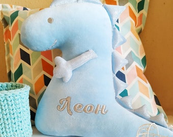 May include: A light blue plush dinosaur toy with a white embroidered name "Leon" on the front. The dinosaur is sitting on a white surface with a colorful patterned pillow behind it.