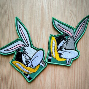 May include: Two embroidered Bugs Bunny patches. Each patch has a green border and shows Bugs Bunny's face in grey, white, black, and yellow. The rabbit is holding a carrot. The patches are on a light brown wooden surface.