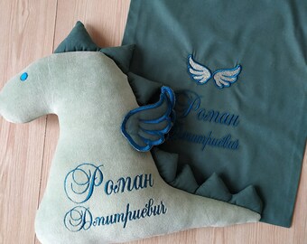 May include: A green plush dragon toy with blue embroidered wings and the name "Roman Dumitru" on it. The dragon is lying on a green fabric bag with a pair of wings and the name "Roman Dumitru" embroidered on it.