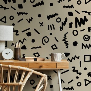 May include: A home office setup featuring a light wood desk, a woven chair, and a beige wall adorned with black abstract shapes. A white lamp, clock, pencils, and a mug are on the desk.