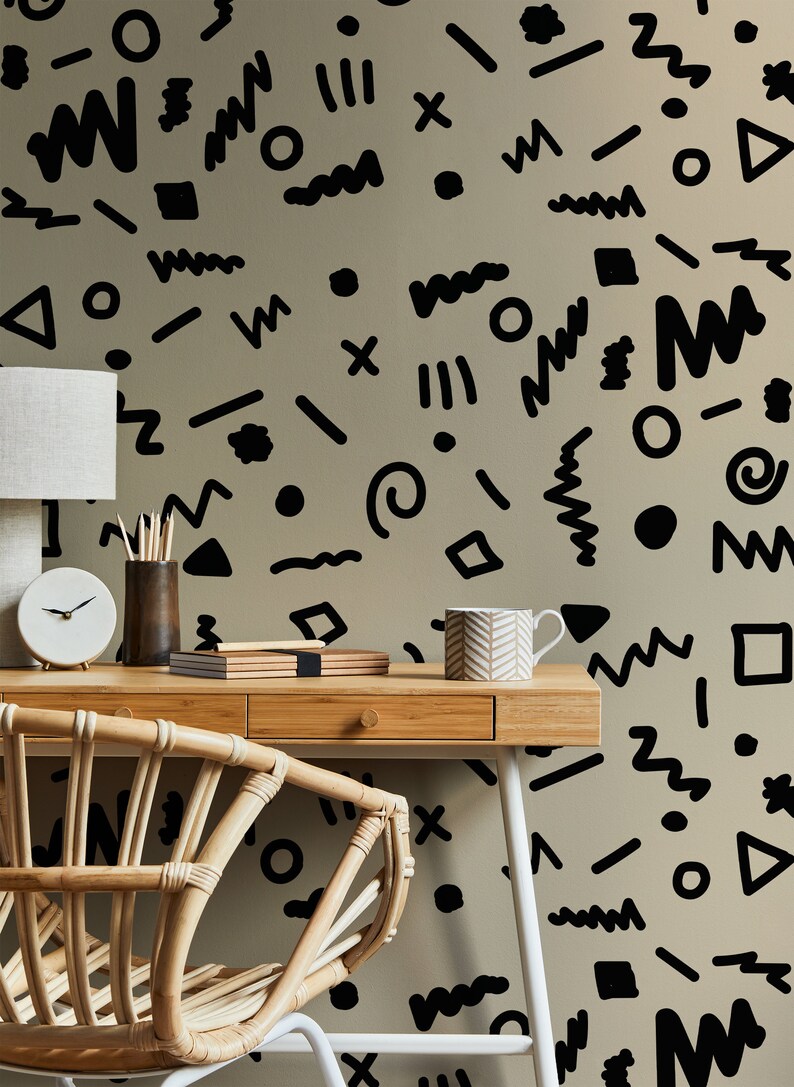 May include: A home office setup featuring a light wood desk, a woven chair, and a beige wall adorned with black abstract shapes. A white lamp, clock, pencils, and a mug are on the desk.