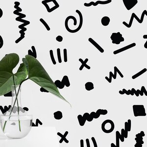May include: A clear glass vase with green leaves, positioned in front of a white wall adorned with black abstract shapes. The shapes include zigzags, circles, and lines, creating a modern graphic design. The vase is on a white surface.