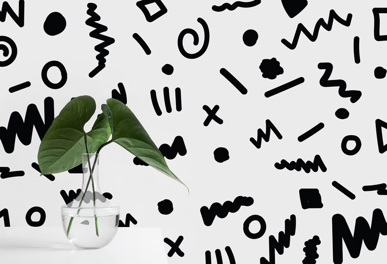 May include: A clear glass vase with green leaves, positioned in front of a white wall adorned with black abstract shapes. The shapes include zigzags, circles, and lines, creating a modern graphic design. The vase is on a white surface.