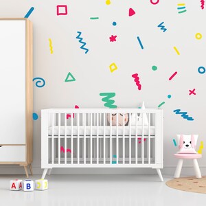 May include: A white cot with a mattress and bedding in a nursery setting. The wall is decorated with colourful shapes. A small pink chair with a dog-shaped cushion sits beside the cot. ABC blocks are on the floor.