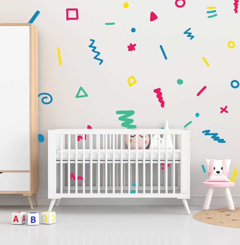May include: A white cot with a mattress and bedding in a nursery setting. The wall is decorated with colourful shapes. A small pink chair with a dog-shaped cushion sits beside the cot. ABC blocks are on the floor.