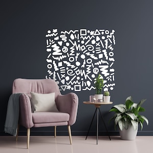 May include: A stylish living room with a pink armchair, a small side table with plants, and a large wall decal. The wall decal is a square of white abstract shapes against a dark grey wall. A grey throw blanket and white cushion are on the chair.