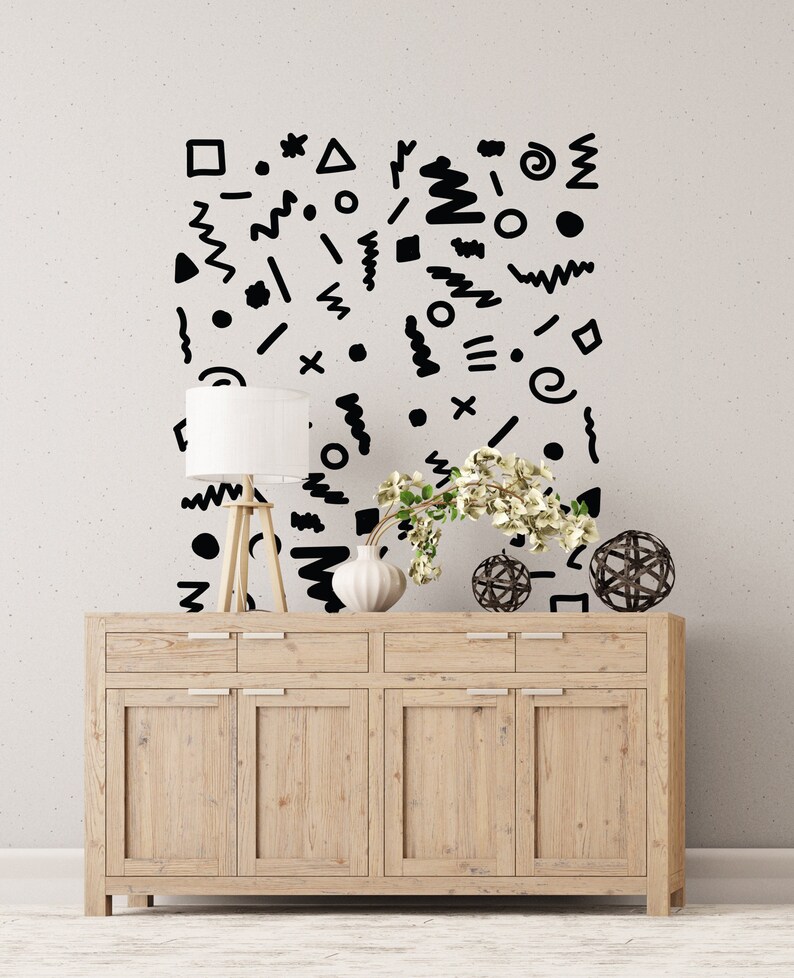 May include: A light wood cabinet with six doors and drawers, a white lamp, a white vase with flowers, and decorative spheres. The wall behind the cabinet has a pattern of black geometric shapes.