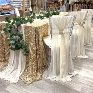 May include: An elegant table setting featuring gold tablecloths and sheer white draping. Clear acrylic chairs are decorated with white fabric and decorative buckles. Greenery and geometric gold accents complete the decor.