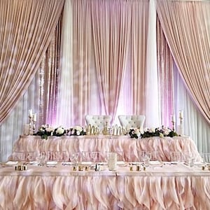 Puede incluir: Una mesa de recepción de boda para los novios con un telón de fondo rosa claro y blanco, una falda de mesa con volantes y dos sillas blancas con letreros de "Mr." y "Mrs.".