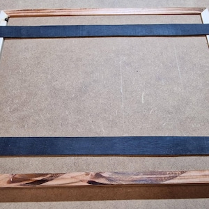 Peut inclure: Un cadre en bois avec des sangles noires attachées à l'intérieur. Le cadre est en bois clair avec une bordure en bois foncé.